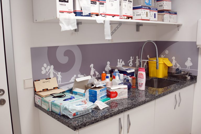 A medical room with a countertop cluttered with various supplies such as gloves, gauze, bottles, and boxes. Above the counter is a shelf with more medical supplies. A sink with a yellow disposal container is also visible.