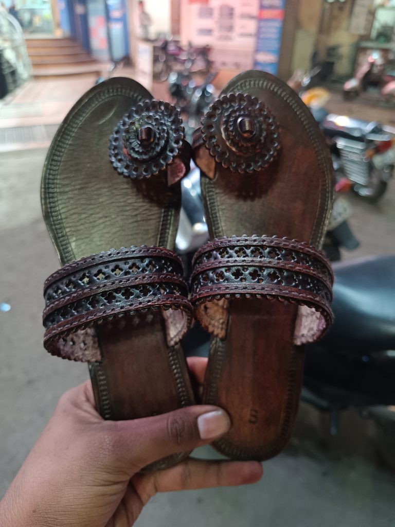 A pair of traditional brown leather sandals with intricate cut-out designs, held in hand, with a background of parked motorcycles and an urban street setting.