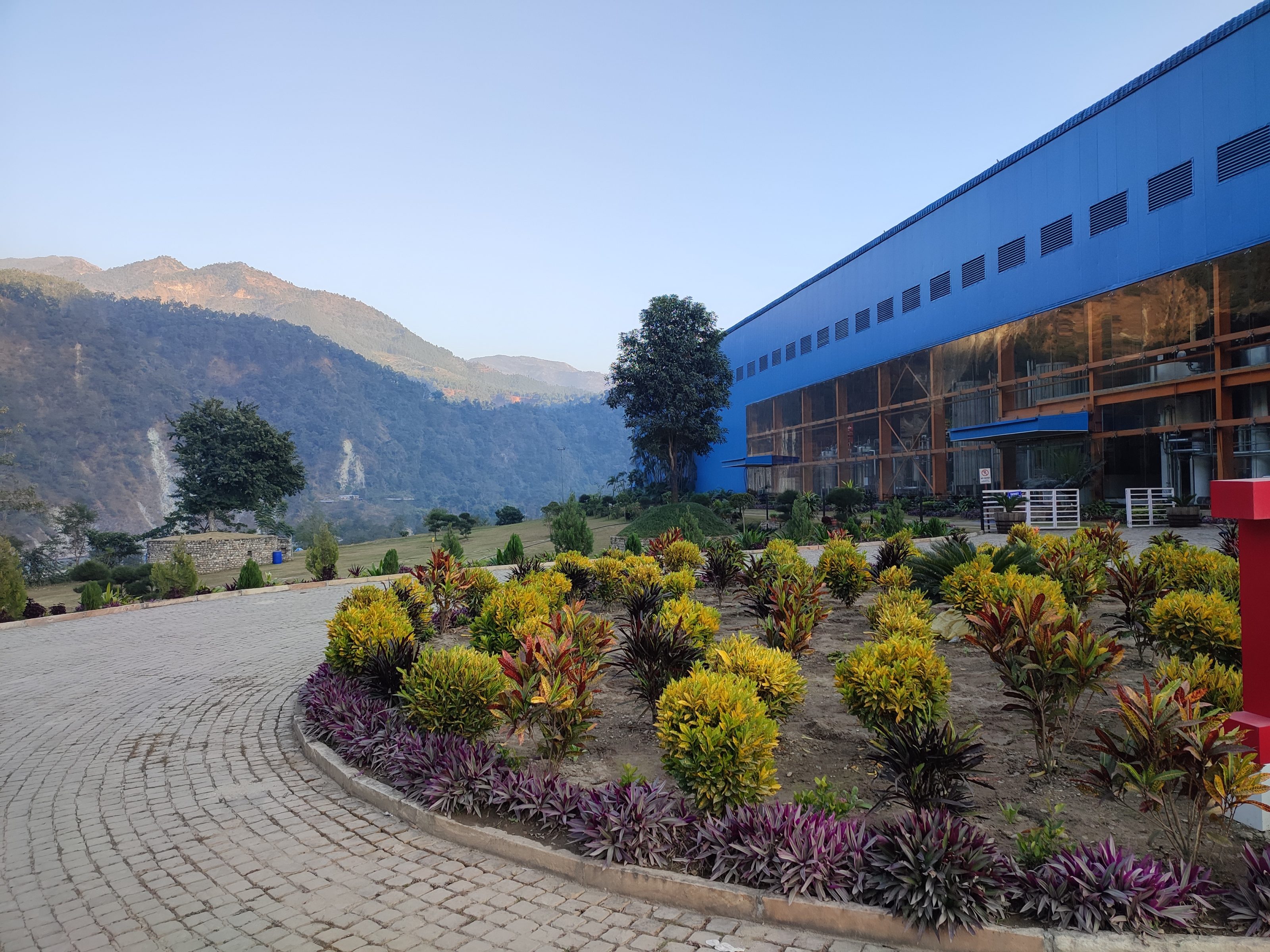 A large industrial building with blue paneling sits adjacent to a landscaped garden featuring colorful shrubs and plants. In the background, there are rolling hills and mountains under a clear sky.