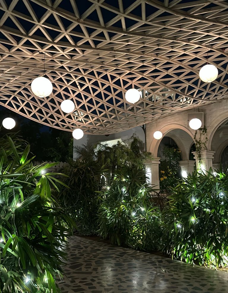 A beautifully lit outdoor space at night featuring a geometric lattice roof with hanging globe lights, lush green plants adorned with string lights, and an arched walkway creating a serene and inviting atmosphere.