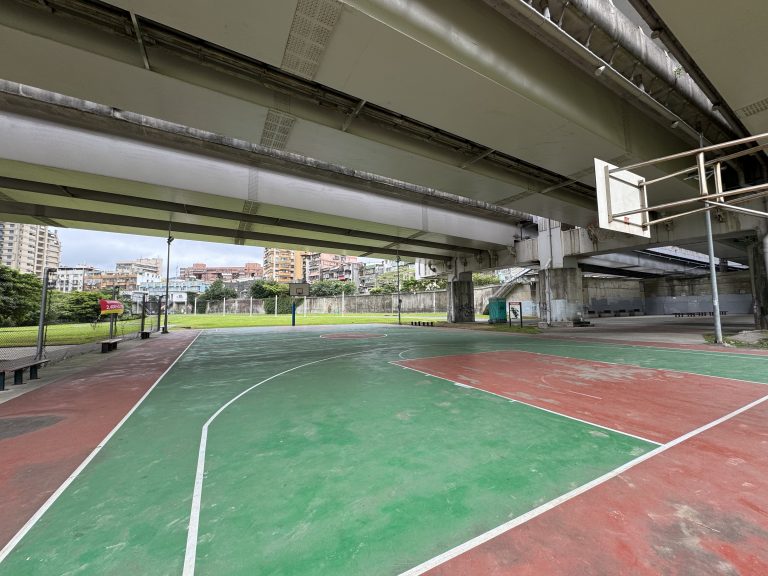 An outdoor basketball court under a large bridge or overpass, with a red and green painted surface. The court is surrounded by benches and a wire fence, and there are residential buildings visible in the background.