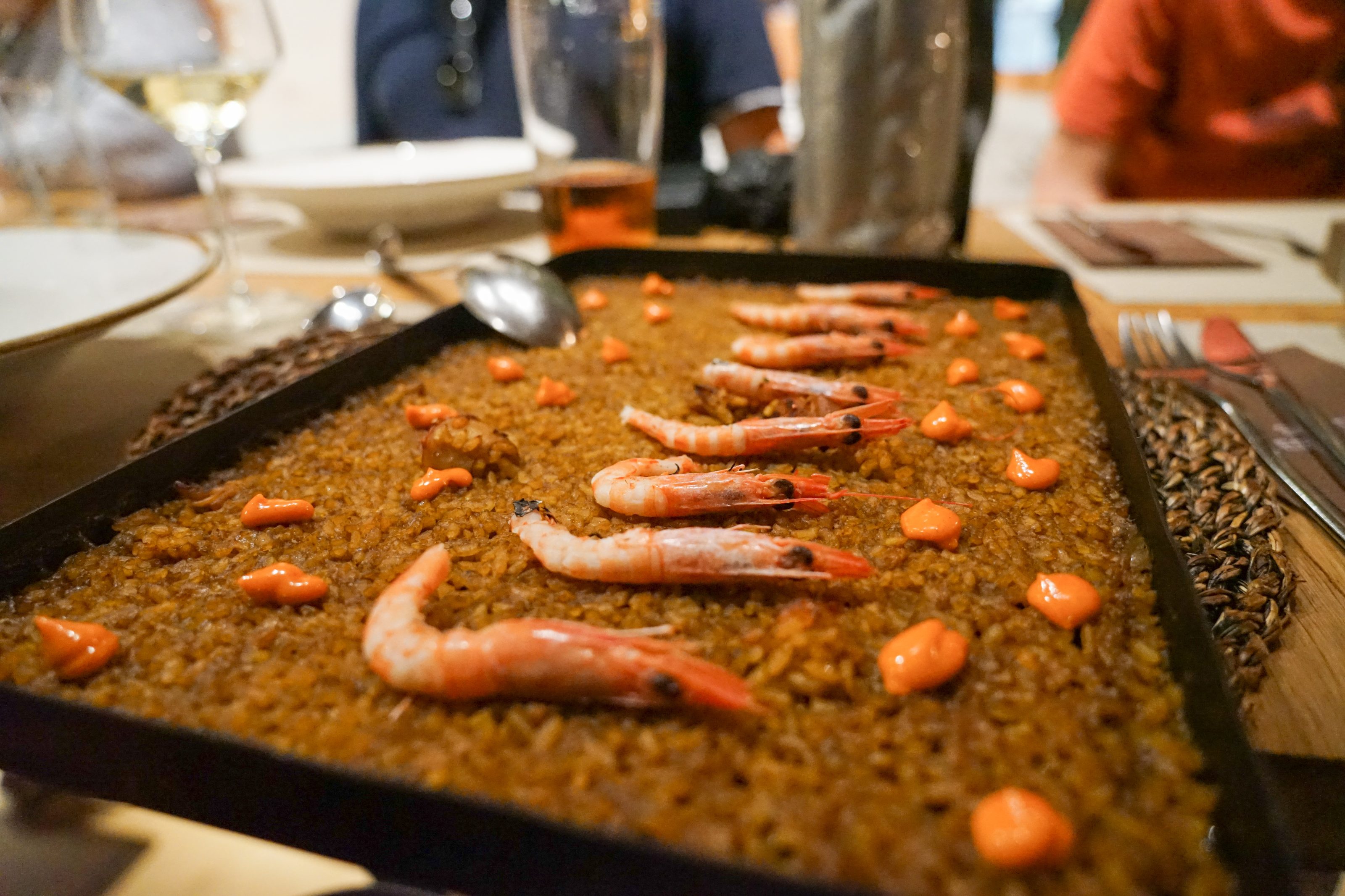 A close-up of a rectangular pan containing a dish of cooked rice topped with whole prawns and small dollops of orange sauce. The pan is set on a dining table, surrounded by glasses, plates, and cutlery, with people seated in the background.