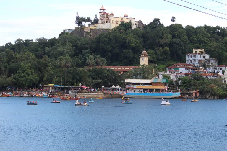 The serene lake is filled with colorful boats, creating a vibrant scene. In the background, lush green hills rise, adorned with the majestic Mount Abu Palace. The city of Mount Abu is visible along the lake, with its charming architecture and vibrant atmosphere.