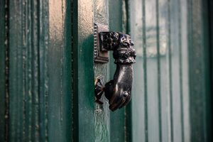 A close-up image of an ornate metal door knocker shaped like a hand holding a ball, mounted on a green wooden door.