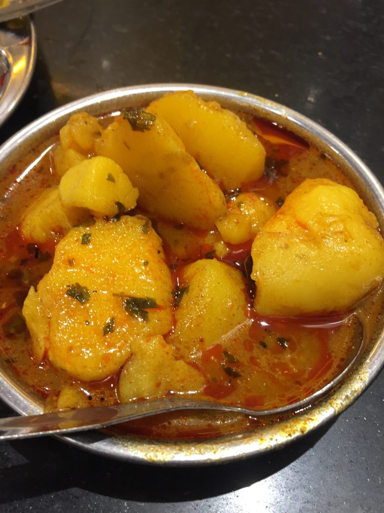 A bowl of spicy Indian potato curry with cooked potato pieces in a rich red-colored gravy, garnished with herbs, placed on a dark surface with a metal spoon inside the bowl.