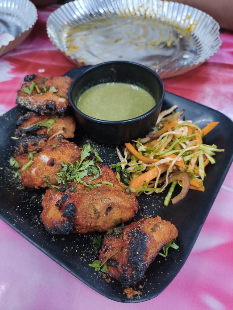 A plate of grilled chicken pieces garnished with chopped herbs, accompanied by a small bowl of green chutney and a side of julienned vegetables.