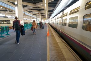 A train station platform with people walking alongside a stationary passenger train. Several individuals carry bags and luggage. The platform has safety barriers and orange cones.