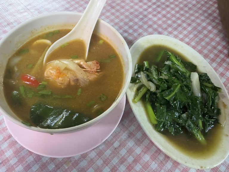 A bowl of spicy Tom Yum soup with shrimp and a side dish of stir-fried Chinese kale served on a pink and white checkered tablecloth.