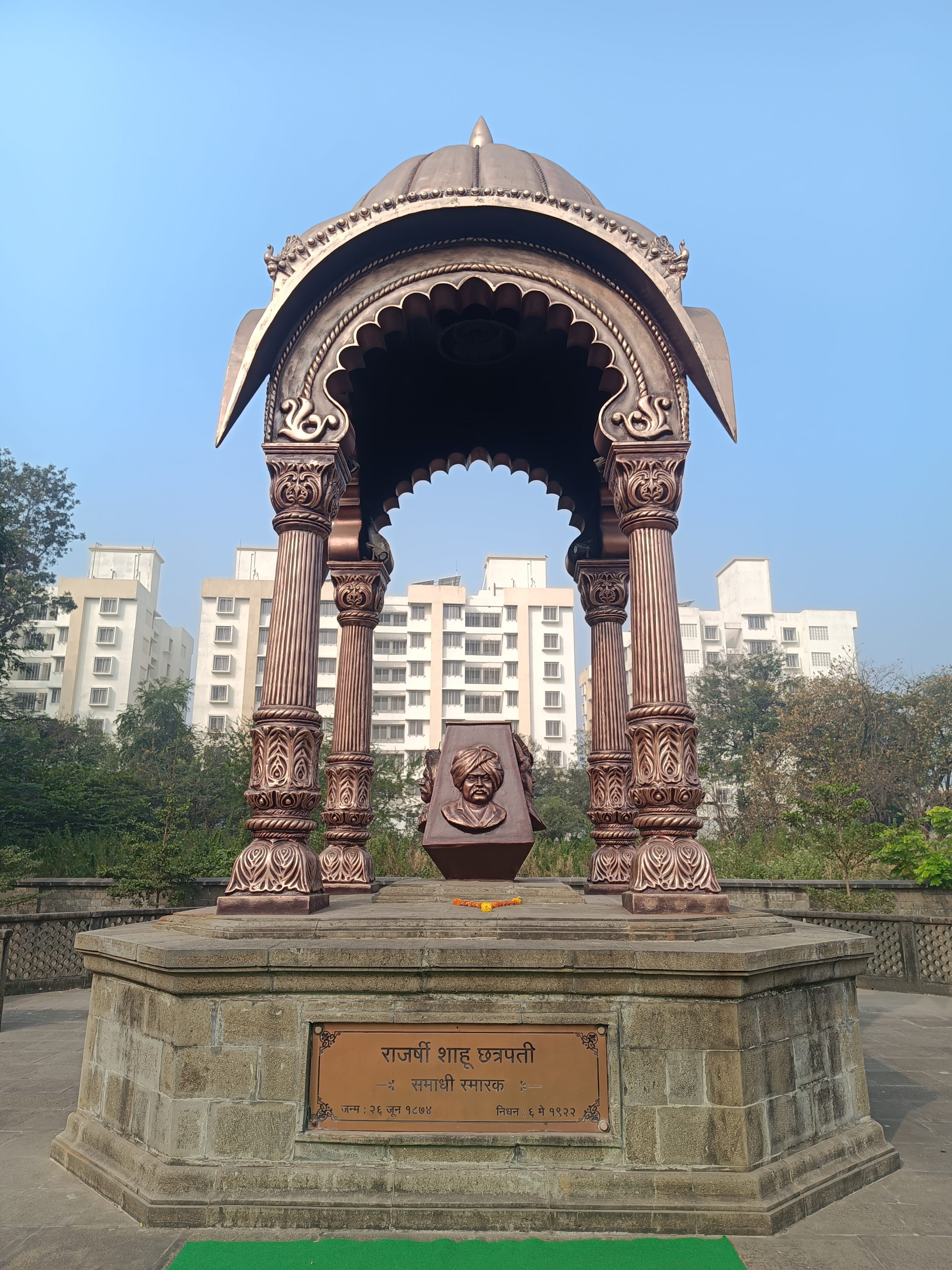 An ornate memorial pavilion dedicated to Rajarshi Shahu Maharaj Samadhi Mandir, Kolhapur. The pavilion features intricately designed pillars and an arched roof, housing a bust on a pedestal. The base of the pavilion includes an engraved plaque with inscriptions. The structure is set against the backdrop of tall residential buildings