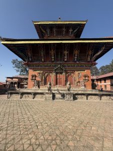 A traditional multi-tiered pagoda-style temple with ornate carvings and vibrant colors, set against a clear blue sky. The foreground features a cobblestone courtyard, and stone sculptures are positioned in front of the temple entrance.