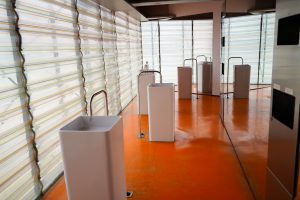 An indoor public space with an orange floor featuring a row of modern rectangular sinks with tall, curved faucets. The walls are made of transparent textured panels allowing natural light to diffuse inside.