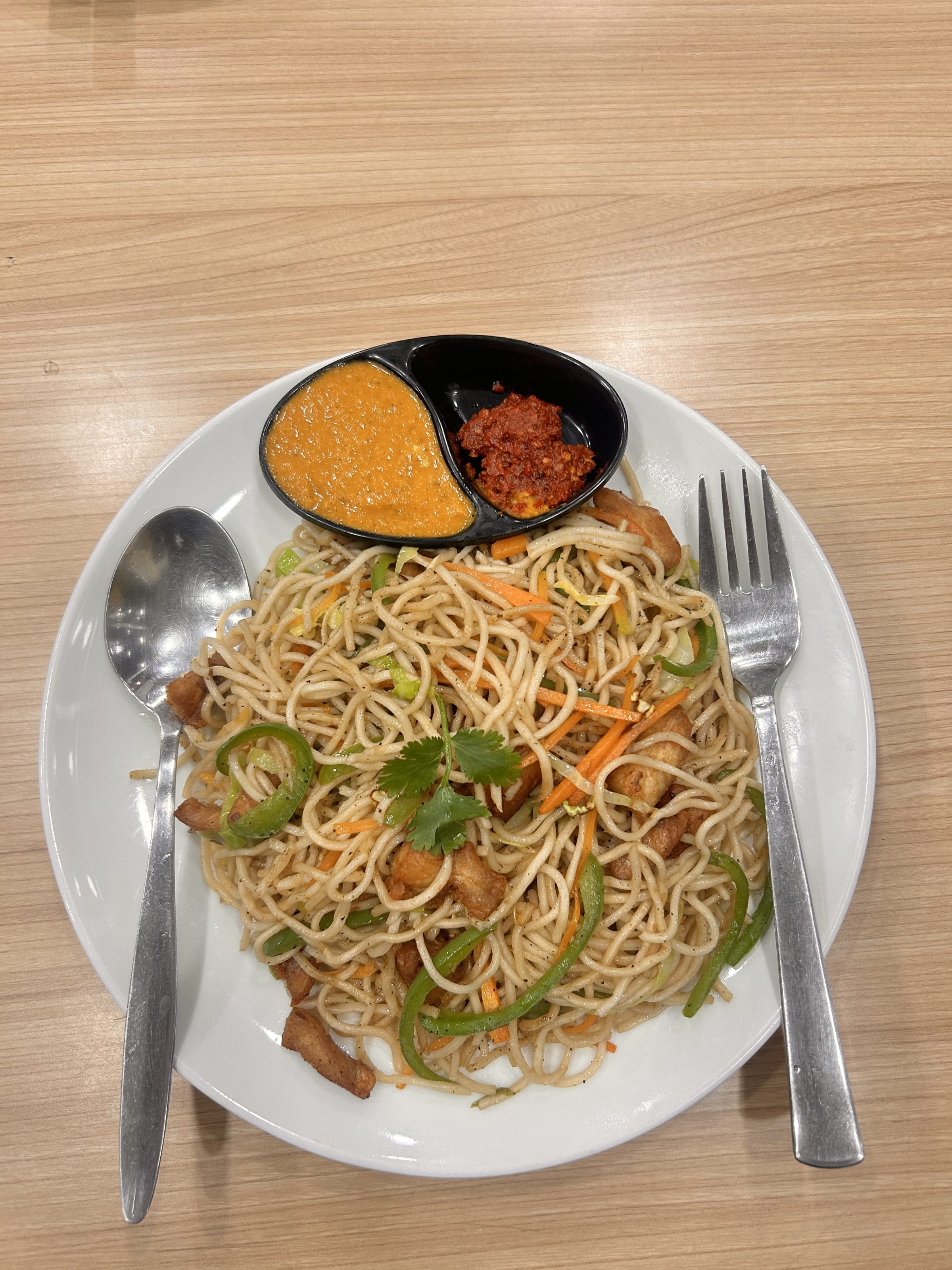 A plate of stir-fried noodles with slices of green bell pepper, carrots, and chicken, garnished with cilantro. Two sauces in a divided dish are placed next to the noodles, and a spoon and fork are arranged on the plate's edges.
