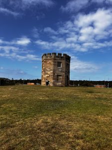 A small, round stone tower with battlements stands on a grassy hill under a partly cloudy blue sky. A person is leaning against the tower, surrounded by a wide open field.