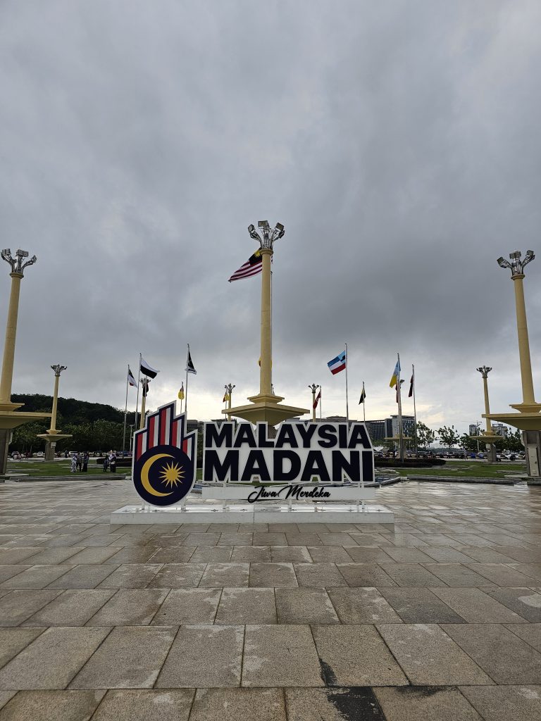 A large sign reading “Malaysia Madani” with a logo featuring the sun and crescent is prominently displayed on a paved area in front of the Prime Minister’s Office, Kuala Lumpur Malaysia. Several flagpoles with international flags surround the area.