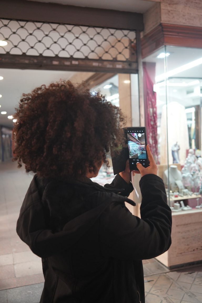 A person with curly hair wearing a black jacket is standing in a shopping area, taking a photo of a store display with a smartphone. The display includes various items like mannequins and jewelry.