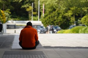 A person wearing an orange hoodie is sitting on the pavement, facing away from the camera, with a blurred parking area and greenery in the background.