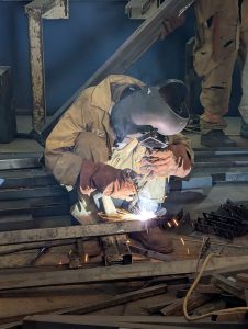 A person wearing protective gear and a welding helmet is crouched down, engaged in welding metal beams for a metal display case.