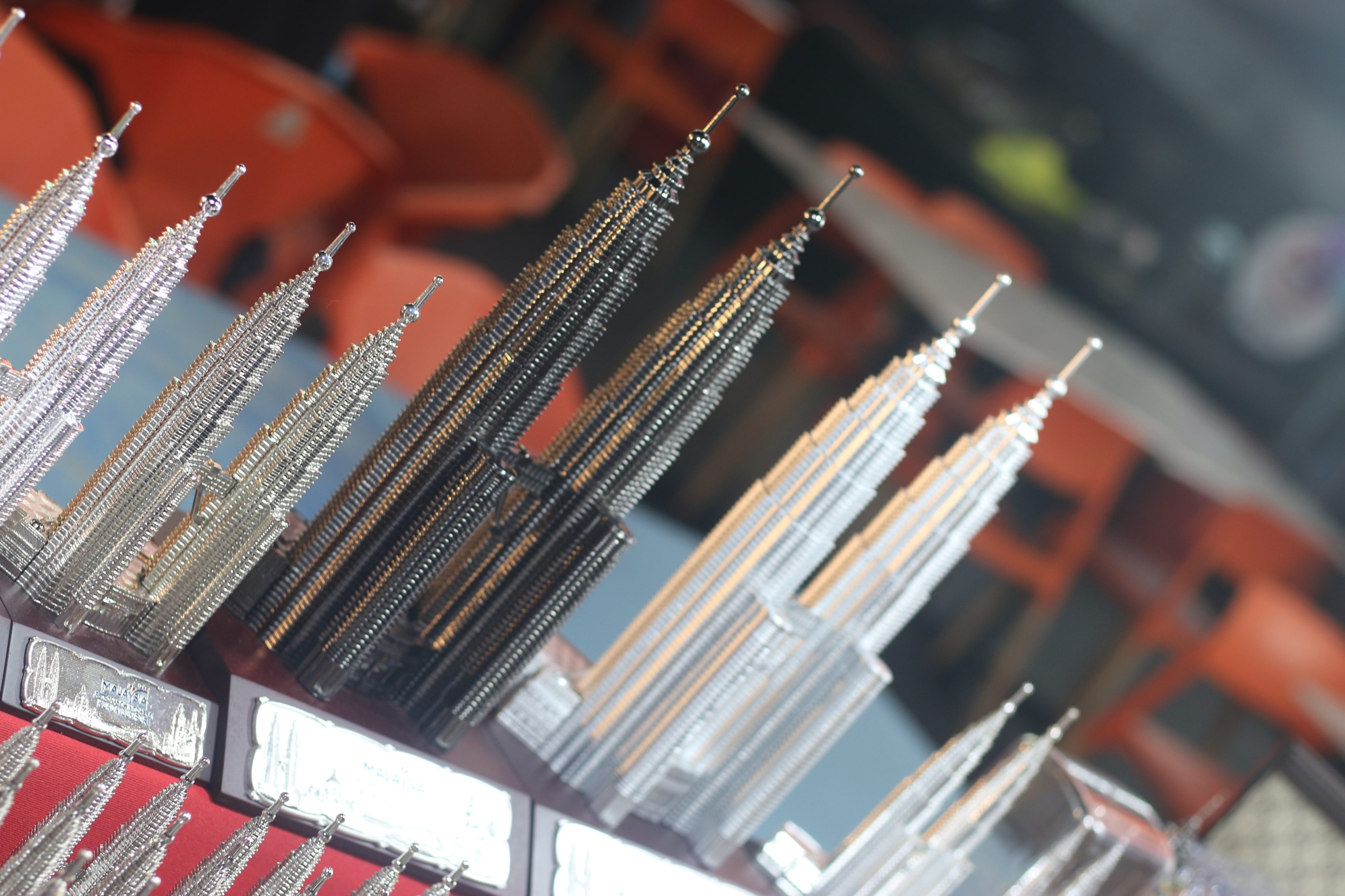 Miniature replicas of the Petronas Twin Towers in silver and black finishes, displayed on a red surface, with blurred background chairs and tables.