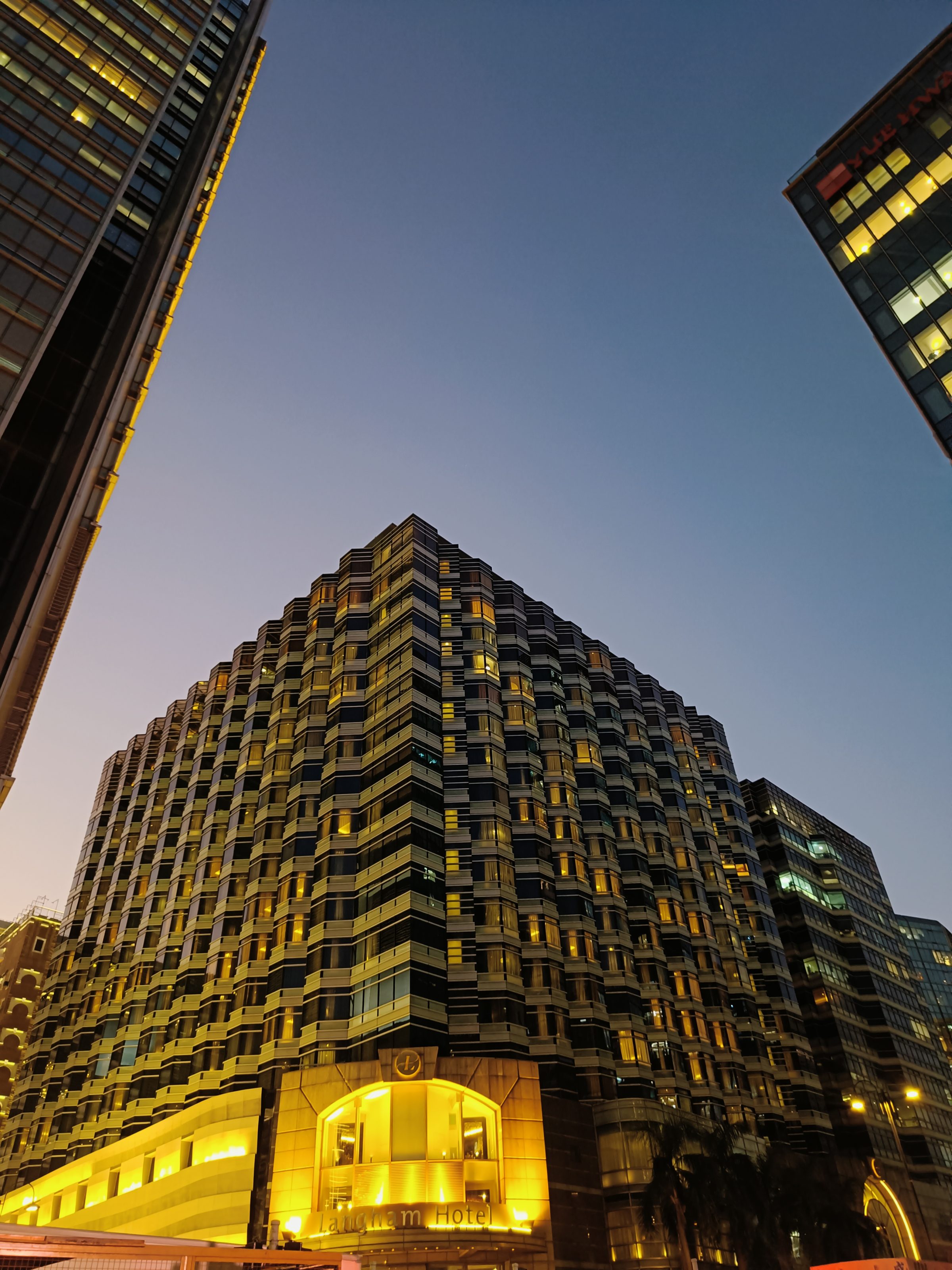A cityscape at dusk featuring a high-rise building with illuminated windows.The sky above is clear, transitioning from a deep blue to a lighter hue as it meets the horizon, bordered by additional tall buildings.