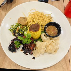 A white and circular plate full of yellow rice, noodles, pickles, chips and chicken pieces. 