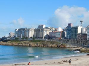  A scenic view of a coastal city with tall buildings lining the shoreline. The foreground features Orzán beach with gentle waves and a few people walking or sitting near the water. A Coru?a, Spain.