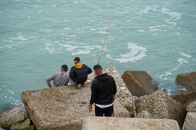 Three people are sitting and standing on large concrete blocks by the sea, with two fishing rods propped up nearby.