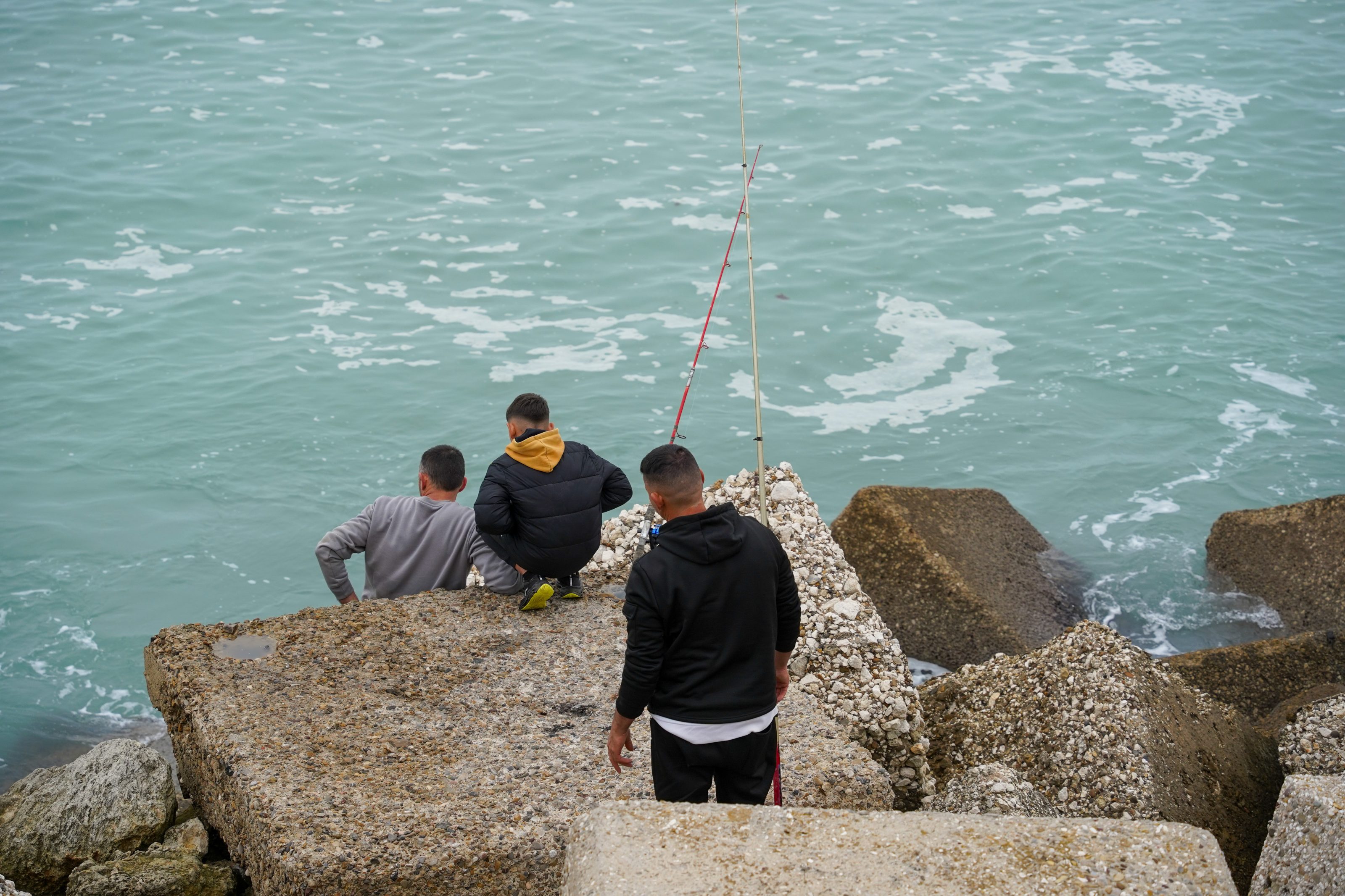 Three people are sitting and standing on large concrete blocks by the sea, with two fishing rods propped up nearby. 