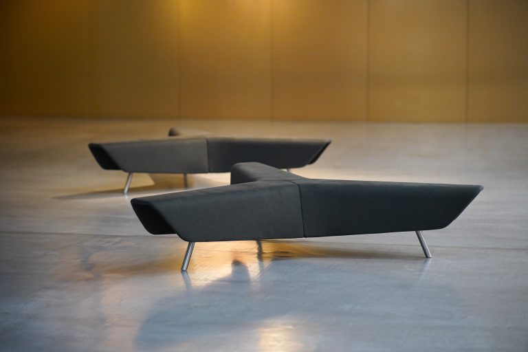 Two modern, angular sofas with a unique geometric design placed on a smooth concrete floor. The lighting casts soft shadows, and the background features a plain, warm-colored wall.