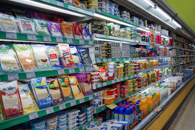 A supermarket aisle with shelves filled with various dairy products, including packaged cheeses, yogurts, and drinks. Bottles of juice and other beverages are also visible on the lower shelves. The aisle is well-lit, showcasing a wide array of colorful products.