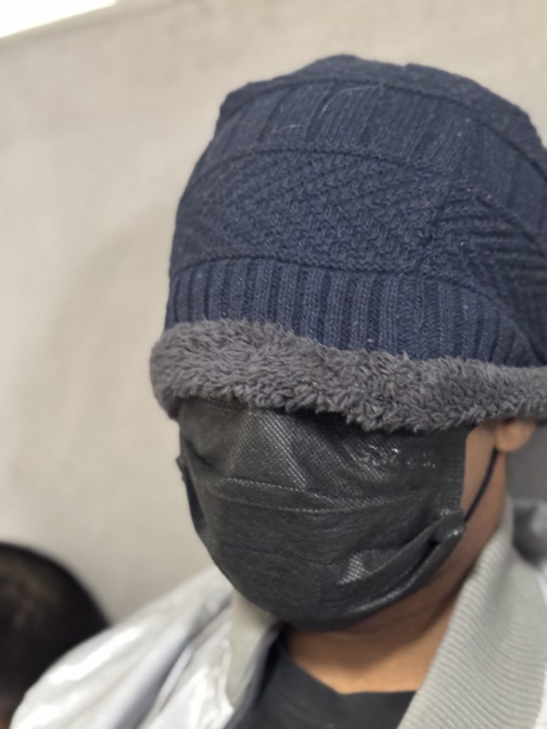 A person wearing a dark knit beanie with a grey fleece band, and a black face mask, partially obscured by shadows.