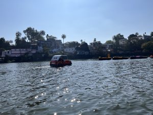 A scenic view of a lake with a pedal boat shaped like a car floating on the water. In the background, there are several colorful pedal boats along the shore, with buildings and trees lining the riverbank