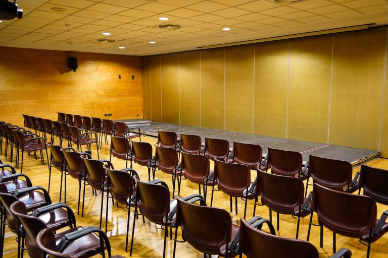 An empty conference room with rows of brown chairs facing a small stage, under an overhead grid-patterned ceiling with recessed lighting. The walls are wooden with a speaker mounted on one side.