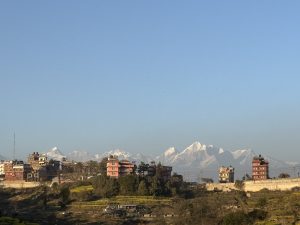 A scenic view of a rural area with scattered multi-story buildings and green fields in the foreground. Snow-capped mountains are visible in the background under a clear blue sky.