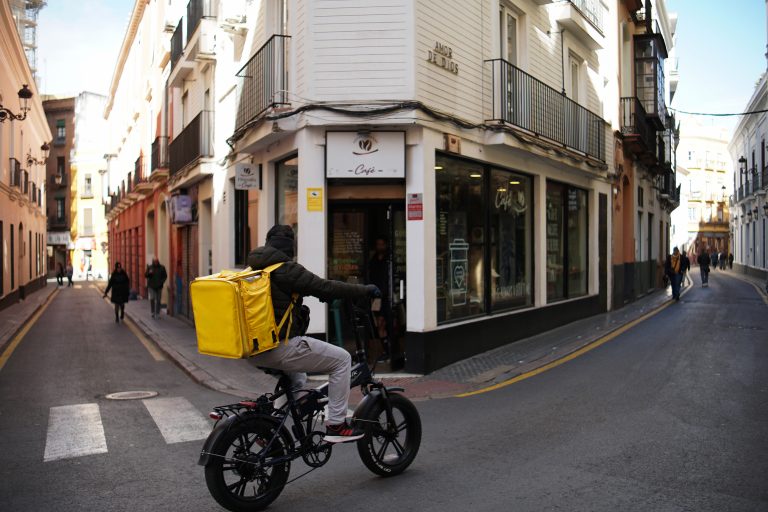 A person wearing a jacket and gloves rides a bicycle with a large yellow delivery backpack through a narrow street lined with old buildings.