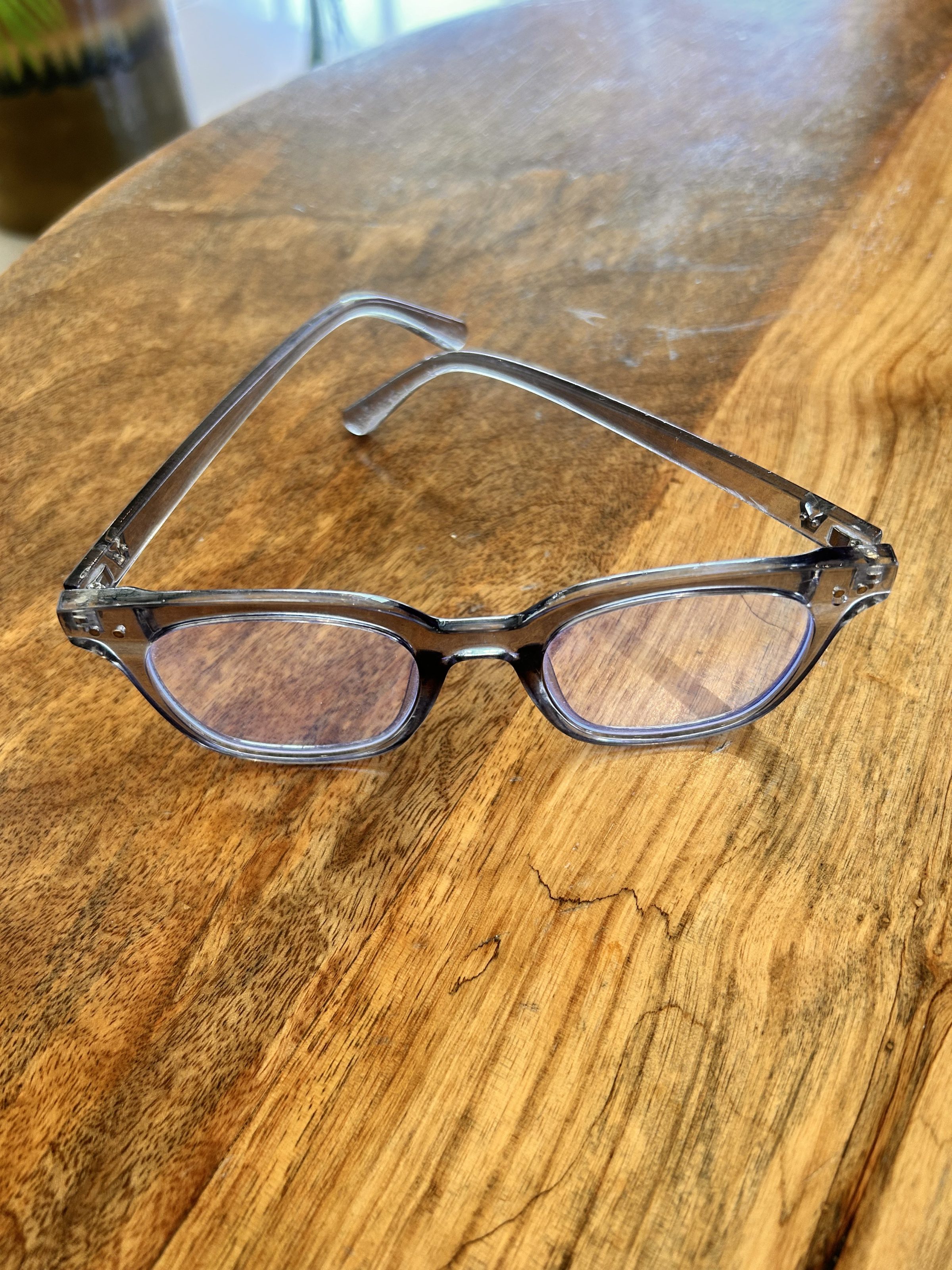 A pair of transparent eyeglasses with blue light filter lenses resting on a wooden table.