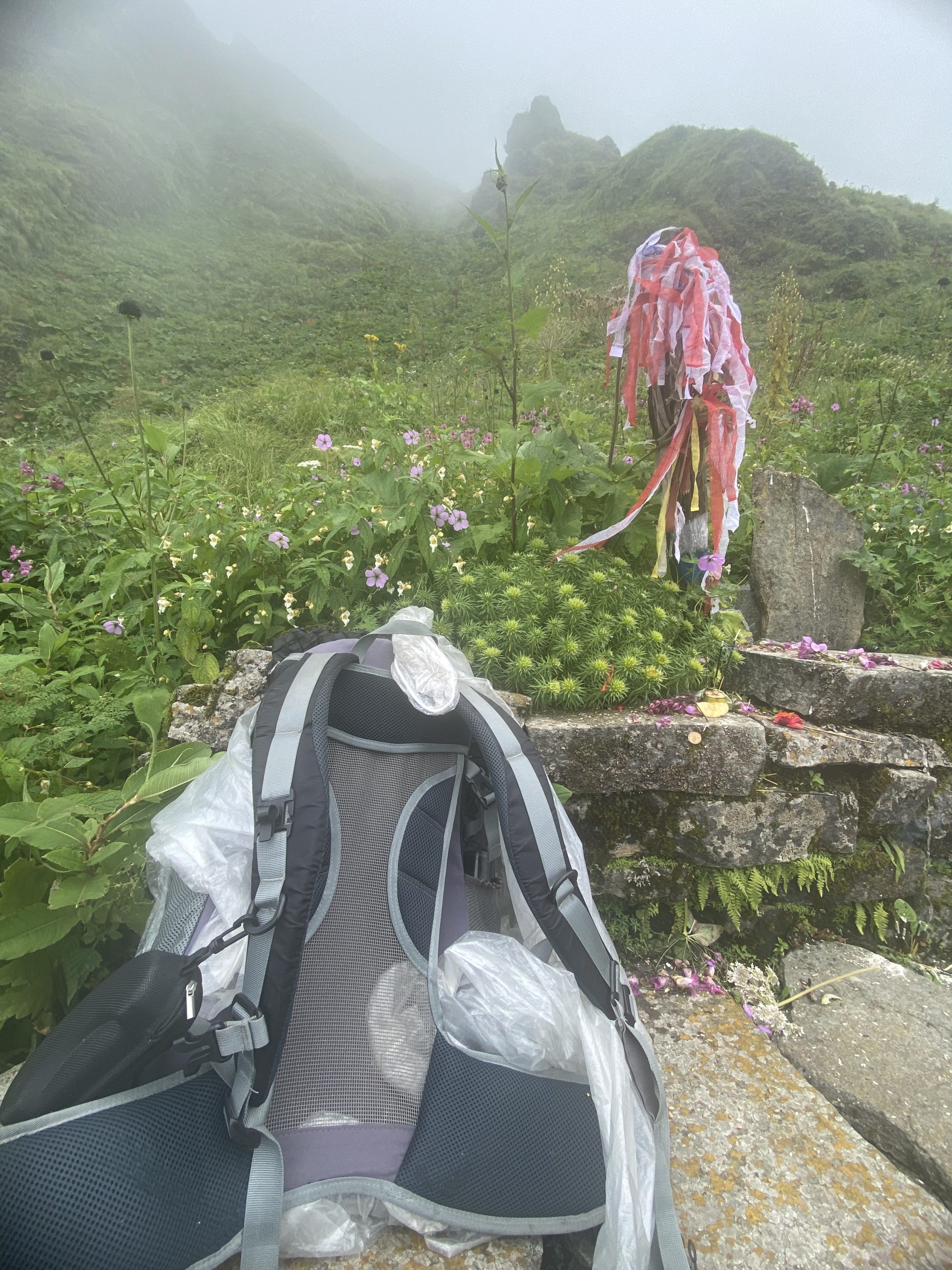 A misty mountainous landscape with lush green vegetation and wildflowers. In the foreground, there is a backpack placed on stone steps, and next to it, a makeshift iron structure with colorful fabric streamers tied around it.