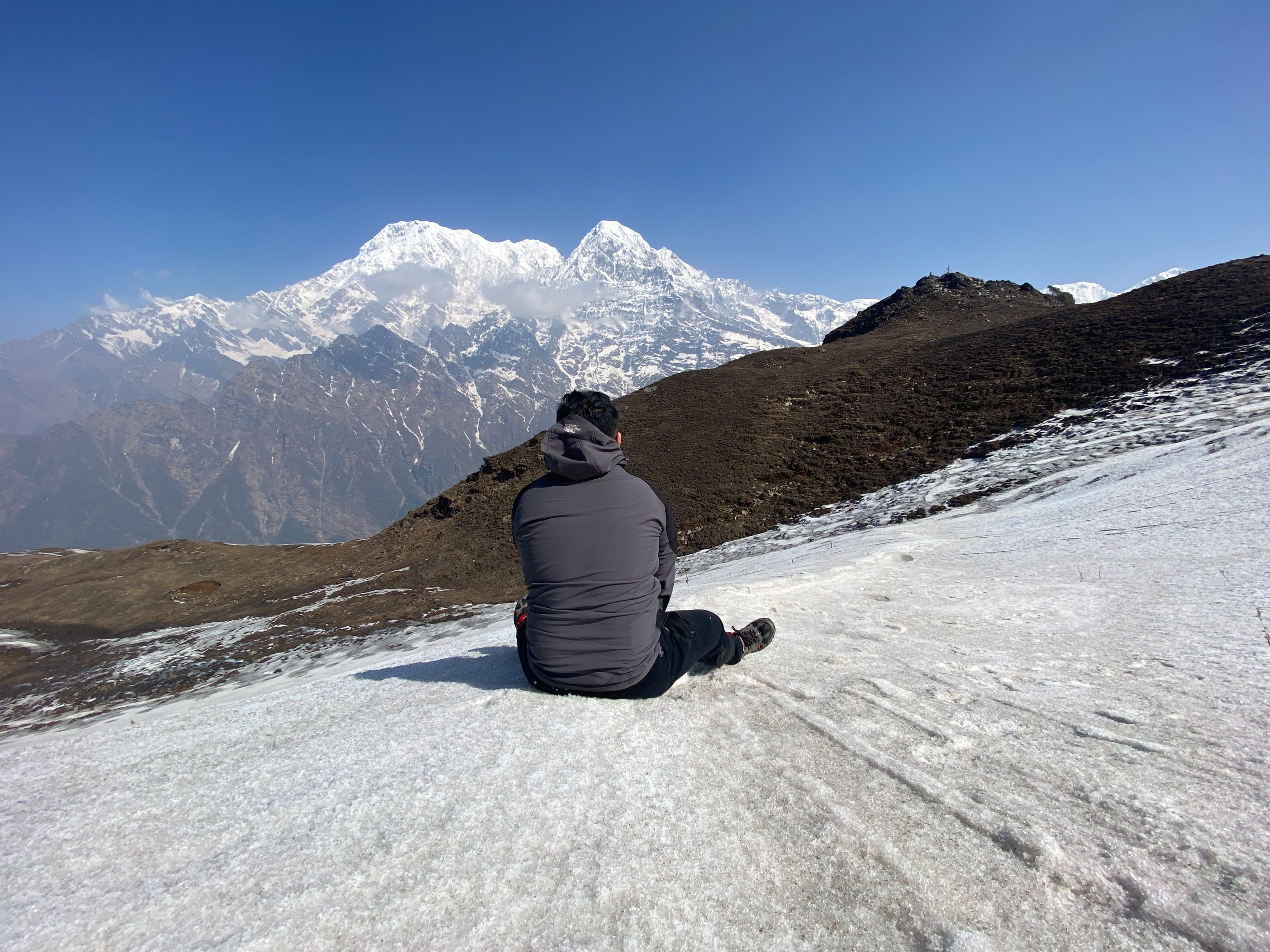 A person sitting on a snowy slope, gazing at a majestic mountain range under a clear blue sky.