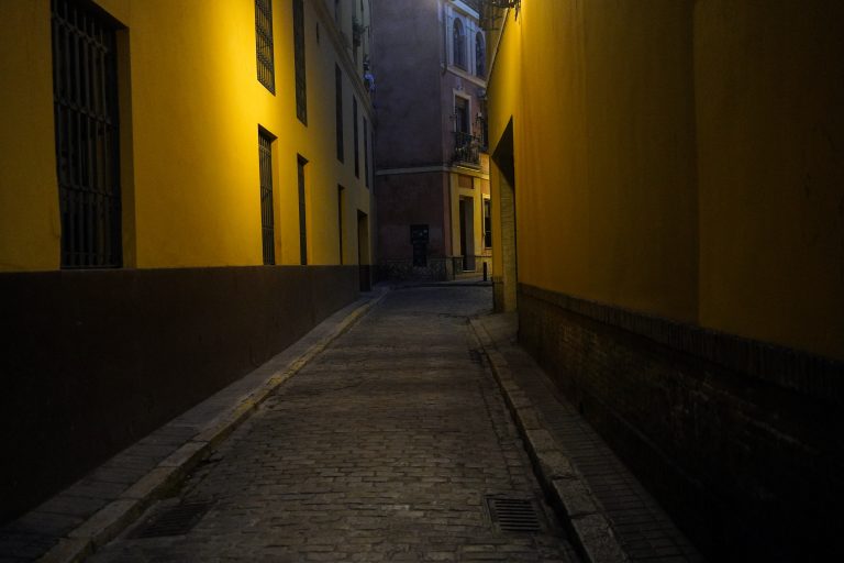 A narrow, dimly lit cobblestone alleyway with yellow walls on either side and a distant view of a building in the background.