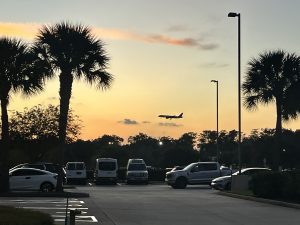 A sunset scene with a silhouette of an airplane flying in the sky, visible beyond a line of palm trees and a parking lot filled with various vehicles. The sky is a gradient of orange and yellow hues with some clouds, creating a tranquil ambiance.
