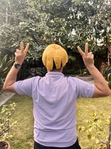 A person standing with their back to the camera in a garden, wearing a yellow citrus peel on their head like a hat, and making peace signs with both hands.