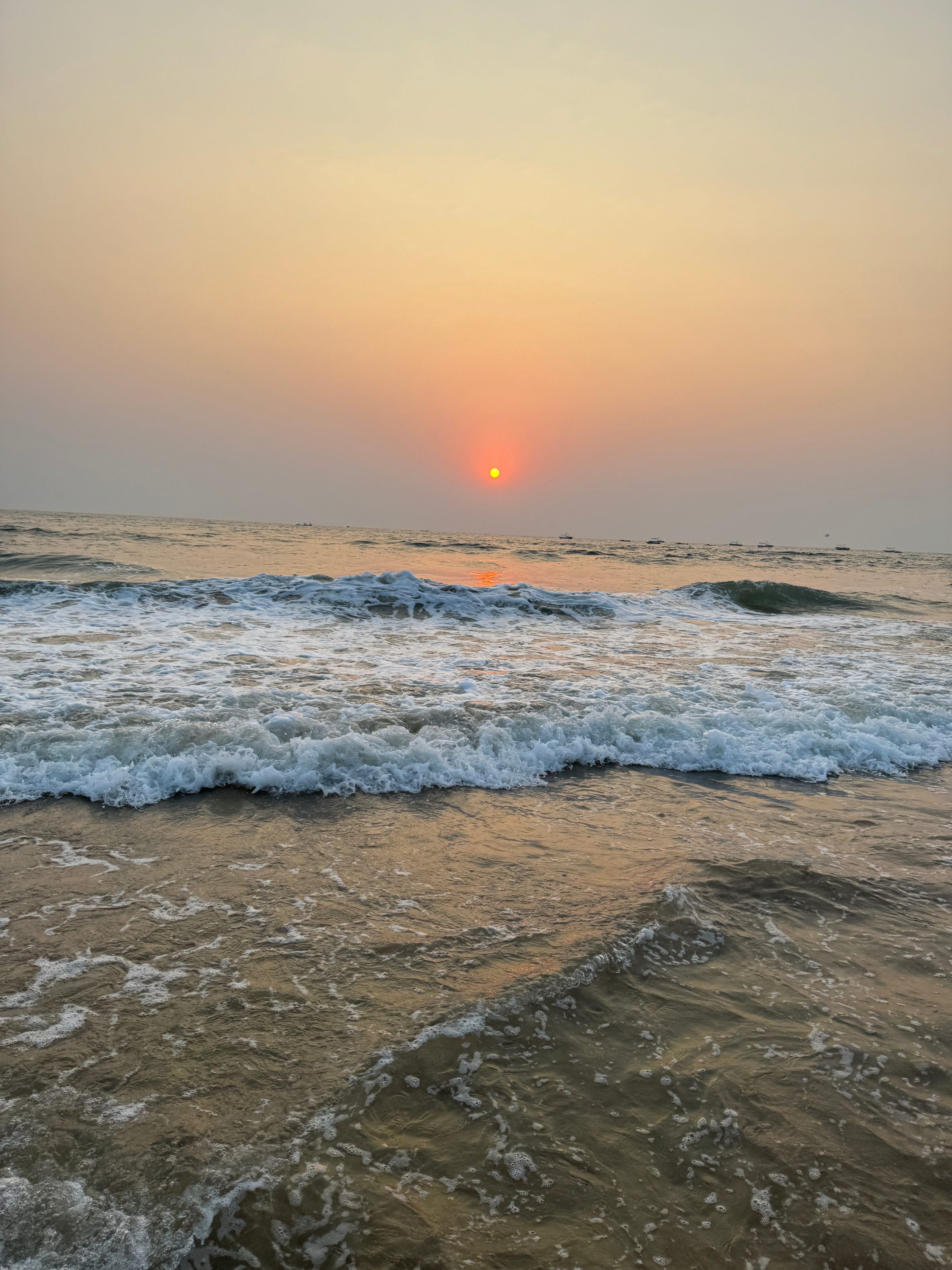 A serene ocean scene at sunset with gentle waves lapping at the shore and a glowing orange sun nearing the horizon, casting a warm reflection on the water.