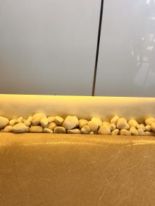 A row of smooth, white pebbles arranged next to a glossy, light-colored stone surface. The pebbles are illuminated with warm yellow lighting, creating a gentle glow against the simple, light gray background.