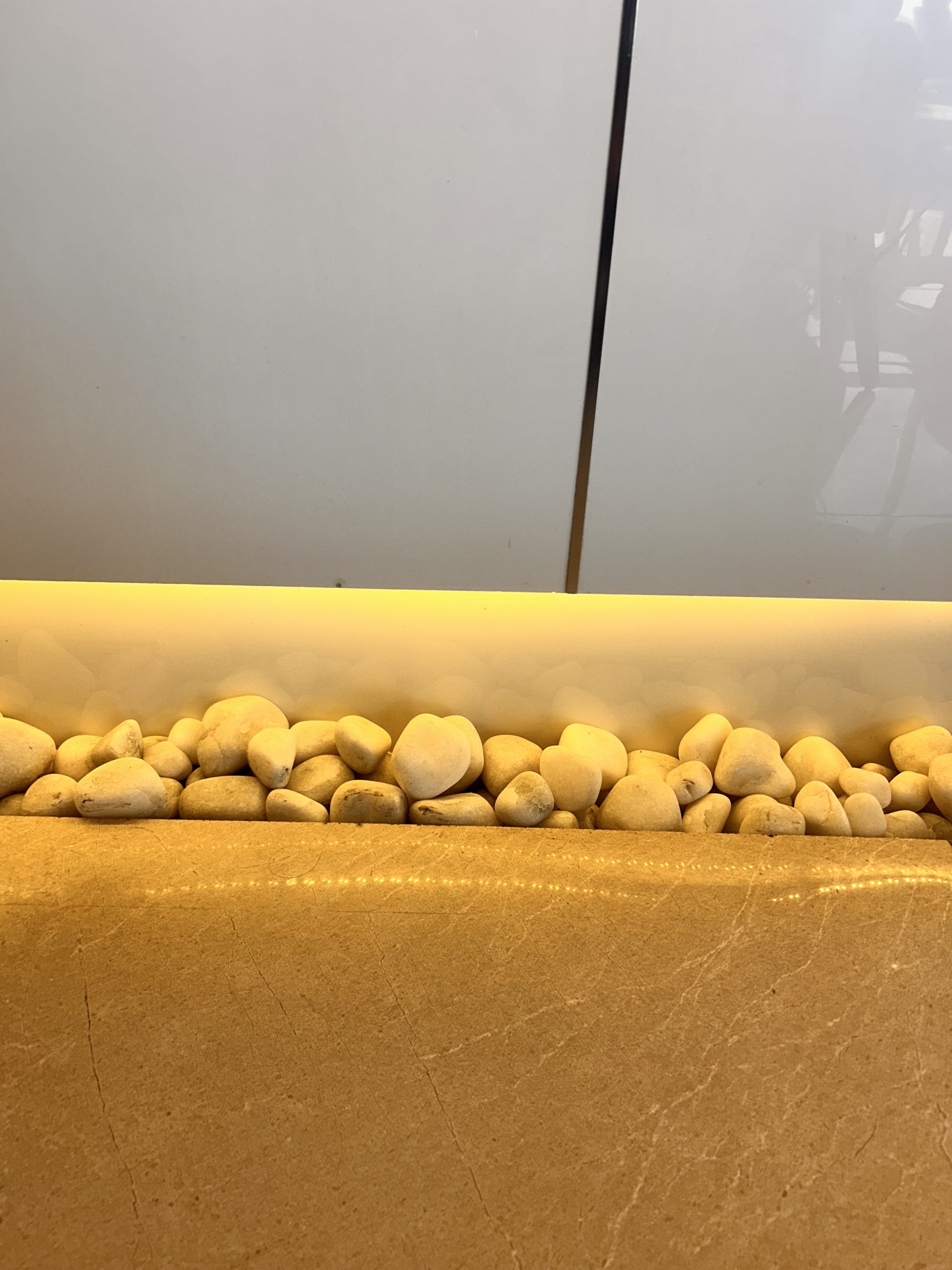 A row of smooth, white pebbles arranged next to a glossy, light-colored stone surface. The pebbles are illuminated with warm yellow lighting, creating a gentle glow against the simple, light gray background.