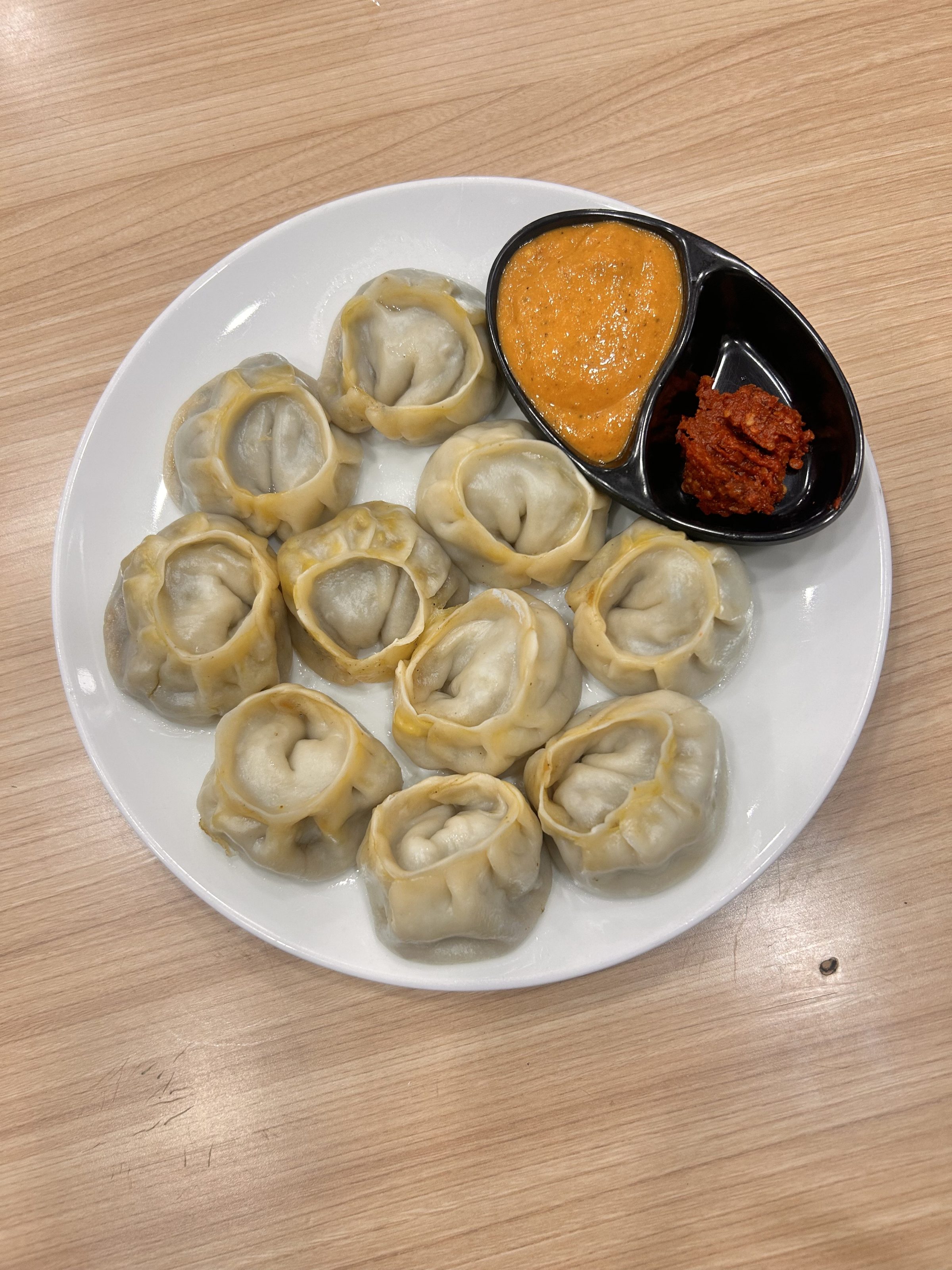 A white plate filled with several steamed dumplings, accompanied by a small black dish containing a bright orange sauce and a red chili sauce, placed on a wooden table.