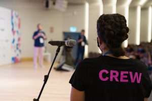 A person wearing a black shirt with "CREW" printed on it in purple stands near a microphone on a stand. The background is a blurred image of a speaker on stage and a few other people in a conference room setting.