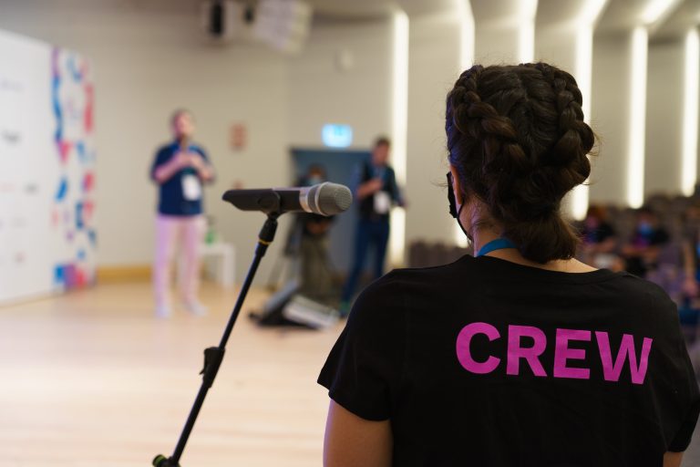 A person wearing a black shirt with “CREW” printed on it in purple stands near a microphone on a stand. The background is a blurred image of a speaker on stage and a few other people in a conference room setting.