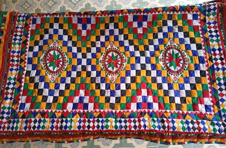 Rajvadi chadar. A colorful, intricately patterned quilt featuring a checkerboard design in vibrant colors such as red, yellow, blue, green, white, and black.