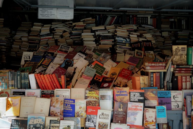 Stacks of various books in different sizes and colors are piled on shelves and tables of a store, with covers facing outward.