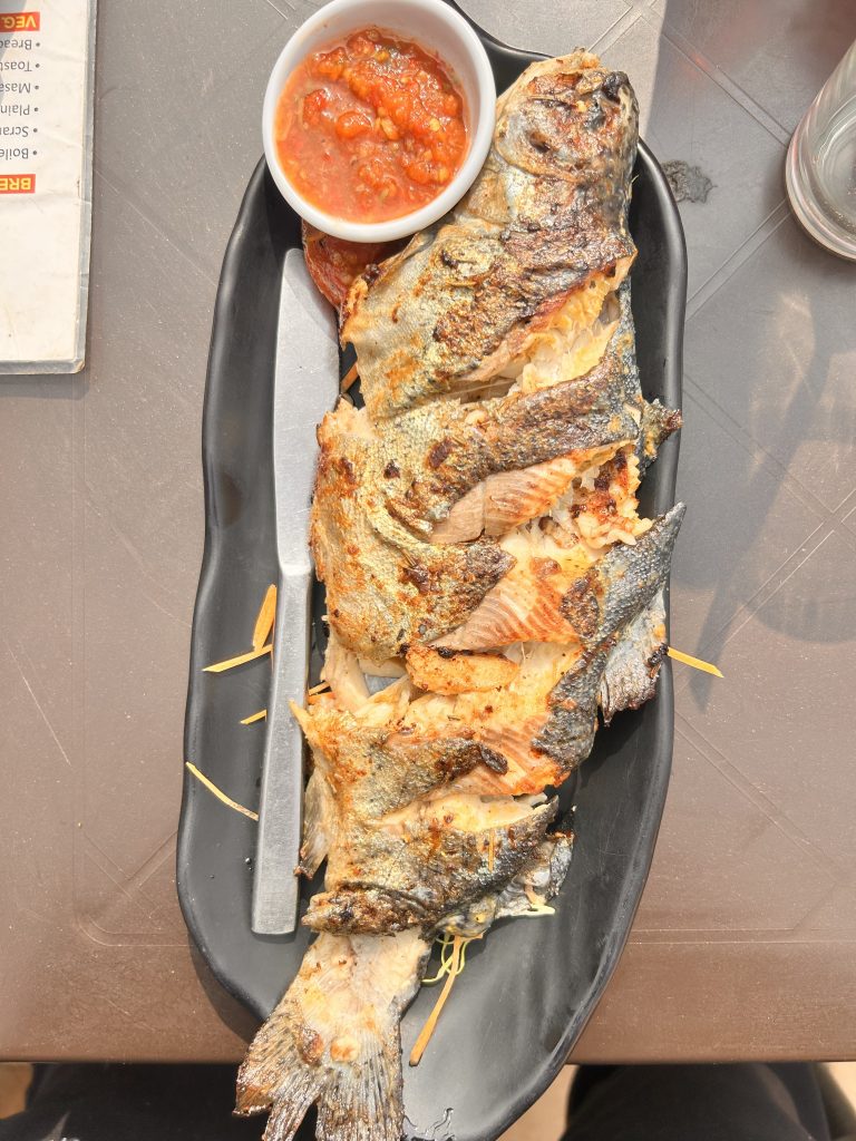 Grilled fish served on a black plate with a side of red sauce in a small bowl. A knife is placed alongside the fish.