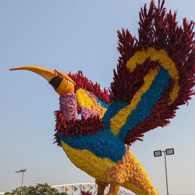 A vibrant floral sculpture of a large bird made with colorful flowers and foliage, featuring shades of red, yellow, blue, and purple, displayed in a garden setting with a clear sky and a bridge in the background.